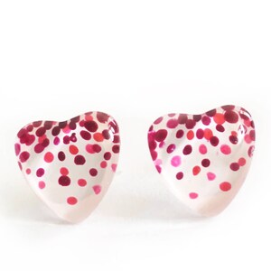 May include: A pair of heart-shaped stud earrings. The clear earrings are decorated with a pattern of pink and red dots. The earrings are set against a white background.