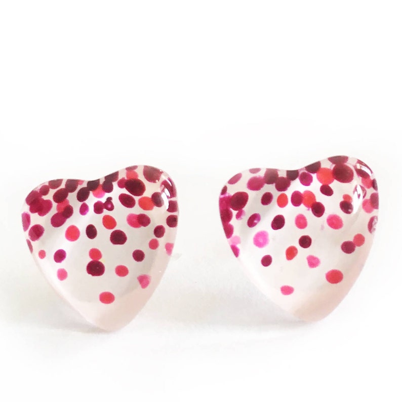 May include: A pair of heart-shaped stud earrings. The clear earrings are decorated with a pattern of pink and red dots. The earrings are set against a white background.