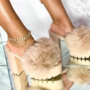 May include: Beige platform sandals with light brown fluffy fur detail. The sandals have a chunky heel and a thick sole. A gold chain anklet is visible on the left foot. The shoes are reflected in a mirrored surface.