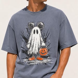 May include: A slate blue short-sleeve t-shirt featuring a graphic print of a character dressed as a ghost with mouse ears. The character is wearing orange shoes and holding an orange jack-o'-lantern bucket. The design has a sketch-like aesthetic.