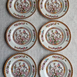 May include: Set of six white ceramic plates with a floral design. The plates feature a brown border with a repeating pattern of small squares. The center of each plate has a floral design with a tree branch with pink and yellow flowers.