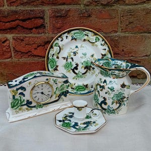 May include: A collection of vintage porcelain tableware featuring a floral design in green, white, and navy blue. The set includes a clock, plate, pitcher, teacup, and saucer, all with gold trim. The clock face has gold hands and Roman numerals.