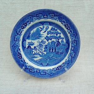 Op de afbeelding: Blauw en wit keramisch bord met een Willow-patroon. Het bord heeft een gedetailleerde landschapsweergave met bruggen, gebouwen en bomen. De rand heeft een decoratieve rand. Het bord wordt op een transparante standaard gepresenteerd.