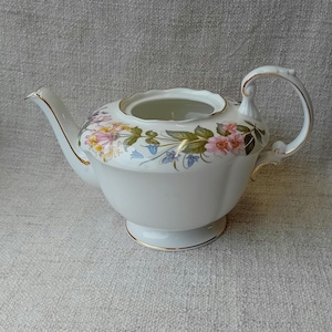 Peut inclure: Théière blanche avec un motif floral dans des tons de rose, jaune et vert. La théière a une bordure dorée autour du bord et de la base.