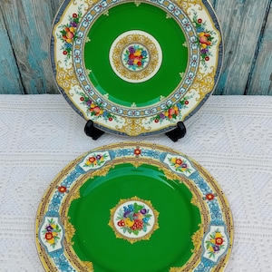 May include: Two green and white ceramic plates with a gold and blue border and a fruit design in the centre. The plates have a white background with a gold and blue border. The centre of the plates has a green background with a fruit design in the centre.