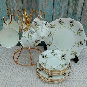 Pode incluir: Um conjunto de chávenas de chá e pires vintage com um padrão floral delicado. As chávenas e os pires são brancos com uma borda dourada e apresentam um desenho de folhas azuis e verdes. O conjunto é apresentado num suporte de metal dourado.