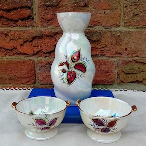 Pode incluir: Um vaso de cerâmica branco perolado e duas tigelas a condizer. O vaso apresenta um design de folhas vermelhas e verdes. As tigelas têm pegas douradas e um padrão de folhas semelhante. O vaso tem cerca de 20 cm de altura.