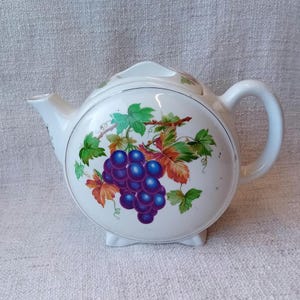 May include: White teapot with a round body and a handle. The teapot is decorated with a cluster of purple grapes on a vine with green leaves and orange accents.