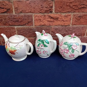 May include: Three ceramic teapots with lids. The teapot on the left has a white background with a floral design in orange, yellow, and green. The other two teapots have a white background with pink flowers and green leaves.