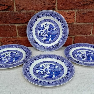 May include: Four blue and white ceramic plates featuring a Willow pattern design. The plates are arranged on a light surface against a red brick wall. The plates have a white centre with a blue landscape scene.