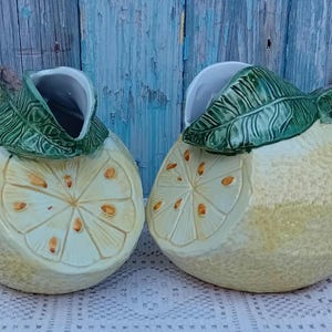 Puede incluir: Dos jarras de cerámica con forma de limón con hojas verdes y un diseño de rodaja de limón amarillo y blanco.