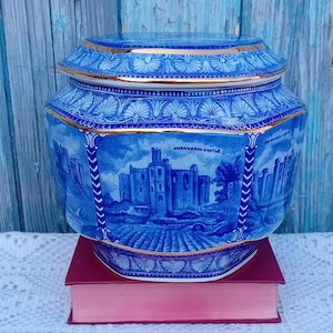 Puede incluir: Un tarro de cerámica azul y dorado con tapa, que representa el Castillo de Warwick. El tarro tiene forma hexagonal y un borde decorativo.
