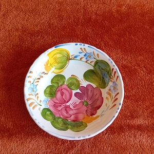 Puede incluir: Un cuenco de cerámica blanco con un diseño floral pintado a mano. Las flores son rosas y moradas con hojas verdes y detalles amarillos y azules. El borde tiene un borde azul y amarillo.