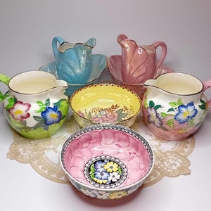 Puede incluir: Colección de jarras y cuencos de cerámica vintage en varios colores pastel. Las jarras tienen asas y diseños florales, mientras que los cuencos presentan patrones intrincados. El conjunto incluye piezas verdes, azules, rosas y amarillas.