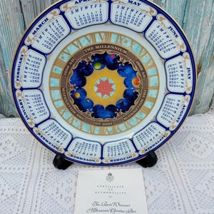May include: A white porcelain plate with a blue and gold border. The plate features a calendar for each month of the year 2000, with a central design depicting the planets and the sun. The text "The Millennium 2000 AD" is written around the edge of the plate.