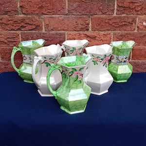 May include: A selection of vintage ceramic jugs in shades of green and white. Some jugs display floral patterns with pink flowers, while others have textured designs. The jugs are arranged on a dark blue surface.