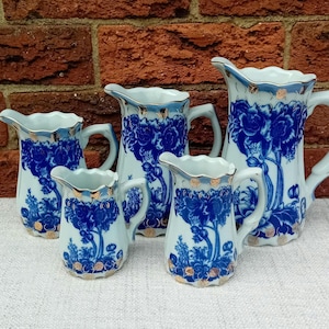 Può includere: Un set di cinque brocche in ceramica bianca con disegni floreali blu e finiture dorate. Le brocche, di varie dimensioni, presentano bordi smerlati e manici curvi. I disegni blu raffigurano alberi e fiori stilizzati.