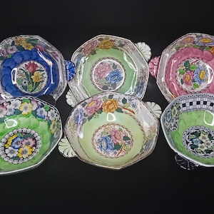 Può includere: Sei ciotole esagonali decorative con disegni floreali. Le ciotole presentano una varietà di colori, tra cui blu, verde e rosa, con finiture dorate e piccole maniglie. Ogni ciotola ha un motivo floreale unico al centro.