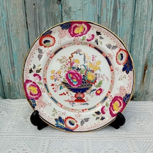May include: A white ceramic plate with a blue and gold rim. The plate features a colorful floral design with a central motif of a vase filled with flowers. The flowers are pink, yellow, red, and blue.