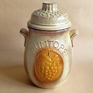 May include: A ceramic jar with a lid, decorated with a brown and white striped pattern and a grapevine design. The jar is labelled "Rumtopf" in brown lettering.