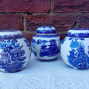 May include: Three blue and white ceramic ginger jars with lids. The jars feature a traditional Asian-inspired design with pagodas, trees, and bridges. The jars are displayed on a white lace tablecloth against a brick background.