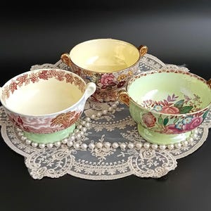 Puede incluir: Tres cuencos de cerámica antiguos con asas, cada uno con un diseño único. Un cuenco tiene una escena pastoral, otro tiene estampados florales y el tercero tiene un interior amarillo. Los cuencos están sobre un tapete de encaje.