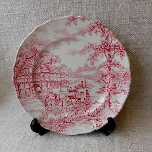 May include: A pink and white ceramic plate featuring a countryside scene with a cottage, a horse-drawn carriage, and trees. The plate has a scalloped edge and is decorated with a pink floral pattern.