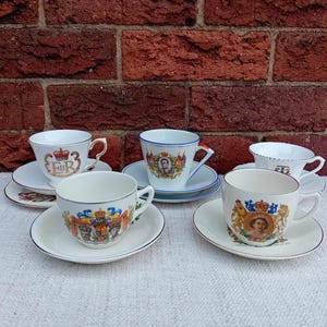 Puede incluir: Conjunto de tazas de té y platillos vintage con diseños de escudos y retratos reales. Las tazas blancas tienen detalles dorados y muestran emblemas y retratos. Los platillos son blancos con diseños a juego. Fondo de ladrillo rojo.