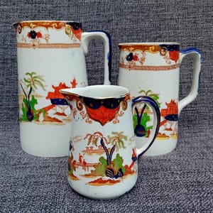 May include: Three vintage ceramic pitchers with a blue and gold floral pattern. The largest pitcher is white with a blue and gold floral pattern. The medium pitcher is white with a blue and gold floral pattern. The smallest pitcher is white with a blue and gold floral pattern.