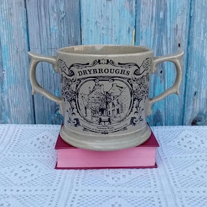 May include: A tan ceramic mug with two handles, featuring a black and white illustration of a building with the text "Drybroughs" and "Tolbooth Wynd Location of Drybroughs first brewery 1750" on the front. The mug also has the years "1750" and "1981" on the sides.