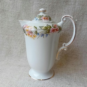 Puede incluir: Cafetera de porcelana blanca con un diseño floral y ribetes dorados. La cafetera tiene una tapa y un asa.