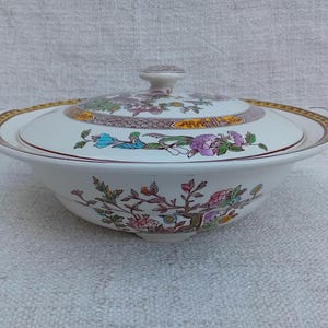 Op de afbeelding: Een witte porseleinen serveerschaal met deksel, met een bloemenpatroon in tinten roze, geel, groen en blauw. De schaal heeft een gouden rand en een decoratief handvat aan elke kant.