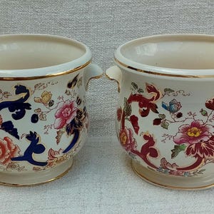 May include: Two white ceramic planters with gold trim and floral designs in shades of blue, red, and green. The planters have two handles on each side.