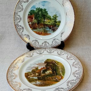 May include: Two white ceramic plates with gold trim and a scenic landscape design. The plates feature a cottage, trees, and a river.