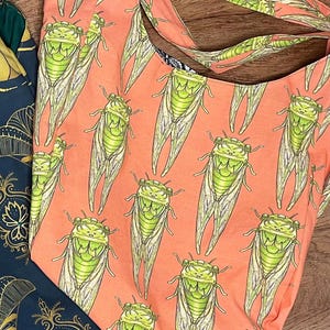 May include: A coral-colored tote bag featuring a repeating pattern of green and white cicada illustrations. The bag has a simple, open design with a matching coral strap. A portion of a dark blue bag with a gold floral design is visible.