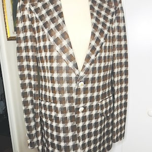 May include: A blazer featuring a brown, black, and white checkered pattern. The garment has a notched lapel, two front flap pockets, and two white buttons on the front closure. Each sleeve cuff is finished with a single white button detail.
