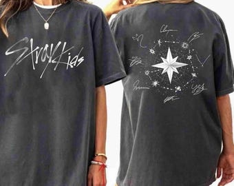 Puede incluir: Una camiseta de manga corta gris carbón con el texto blanco Stray Kids en la parte delantera. La parte trasera muestra un gráfico blanco de una estrella central rodeada de líneas celestiales, estrellas pequeñas y firmas manuscritas en círculo.