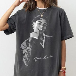 May include: Charcoal grey t-shirt with a black and white portrait of a smiling person. The name "Malachi Burton" is written in a handwritten style. The shirt has short sleeves and a loose fit.