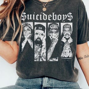 Op de afbeelding: Donkergrijs t-shirt met de bandnaam "Suicideboys$" in witte gotische letters. Het shirt toont een zwart-wit afbeelding van vier bandleden, met het woord "Staff" en het nummer "59" zichtbaar. Het t-shirt heeft een vintage, versleten uitstraling.