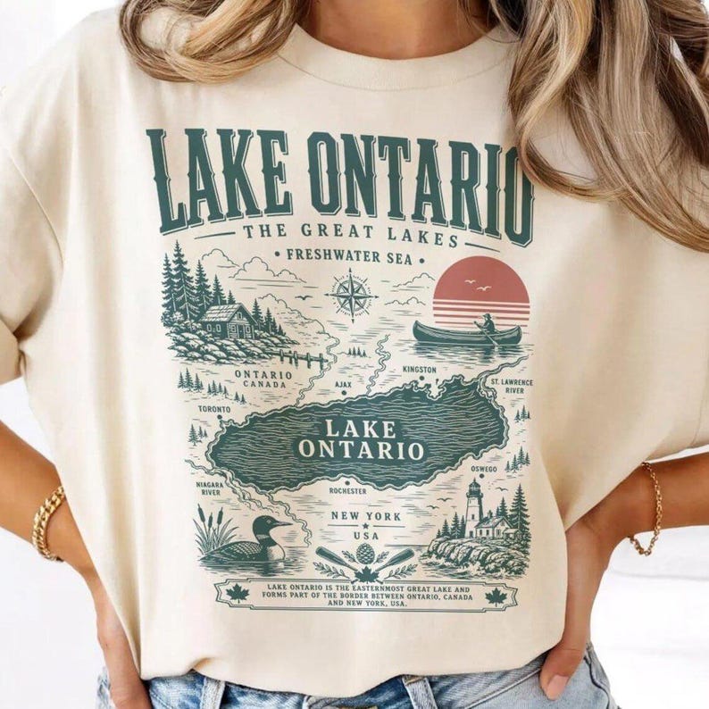 May include: A cream-colored t-shirt featuring a dark green graphic print of a map of Lake Ontario. The design includes the text Lake Ontario, The Great Lakes, Freshwater Sea, and locations like Toronto, Ajax, Kingston, and Rochester, with a canoe.