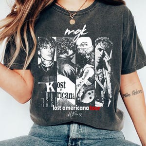 May include: Dark grey t-shirt with a black and white graphic of a musician in four panels. The text "mgk" is above the panels, and "Kost Americana" and "lost americana tour" are below. The shirt has a vintage, worn appearance.