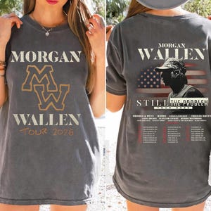May include: Dark grey t-shirt with the text "MORGAN WALLEN TOUR 2026" on the front, and a gold "MW" logo. The back features "MORGAN WALLEN STILL THE PROBLEM TOUR 2026" with a silhouette and American flag design.
