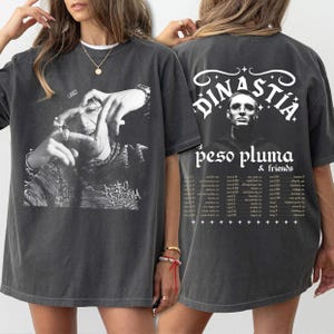 Könnte beinhalten: Dunkelgraues T-Shirt mit einem Schwarz-Weiß-Bild auf der Vorderseite. Auf der Rückseite des Shirts steht der Text "DINASTÍA" über einem Porträt und die Worte "peso pluma & friends" mit einer Liste von Tourdaten.