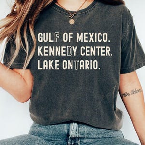 May include: A charcoal grey short-sleeve t-shirt with a distressed white graphic print. The text reads: GULF OF MEXICO. KENNEDY CENTER. LAKE ONTARIO. The letters F, D, and T are stylized with a vertical line through the center of each character.