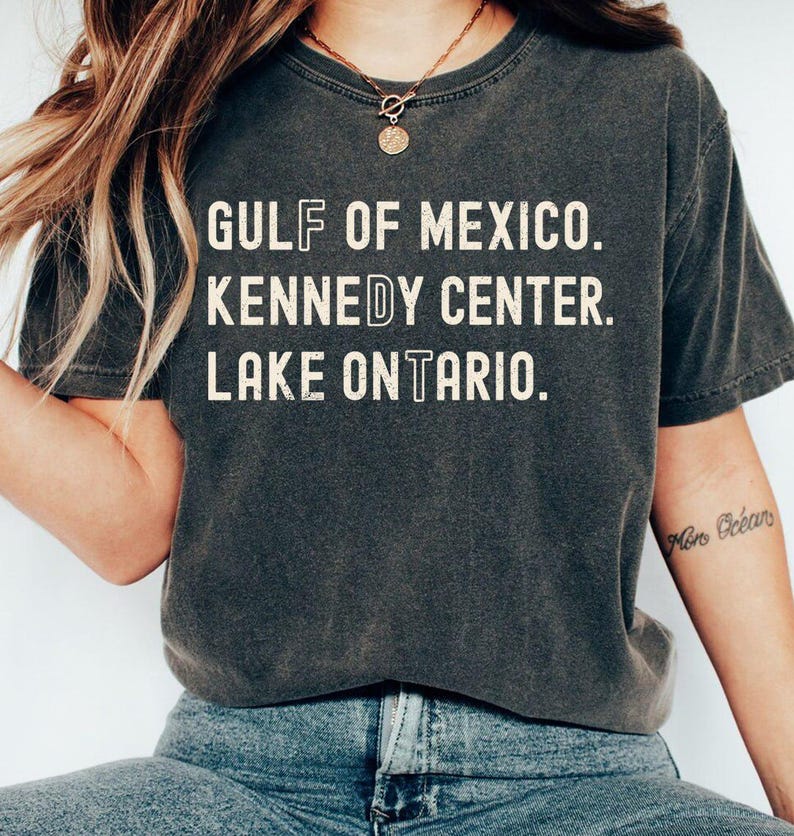 May include: A charcoal grey short-sleeve t-shirt with a distressed white graphic print. The text reads: GULF OF MEXICO. KENNEDY CENTER. LAKE ONTARIO. The letters F, D, and T are stylized with a vertical line through the center of each character.