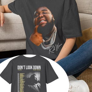 Może przedstawiać: Ciemnoszary t-shirt z krótkim rękawem z portretem Rod Wave z łańcuchem z przodu i napisem Rod Wave. Z tyłu znajduje się napis DON'T LOOK DOWN TOUR ROD WAVE, czarno-biały portret oraz lista dat trasy koncertowej.
