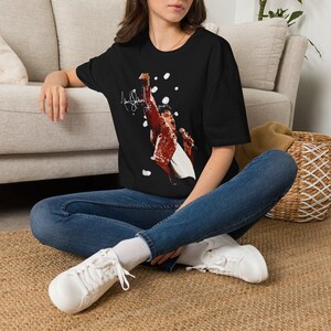 May include: A black short-sleeve t-shirt featuring a graphic print of a singer in a red jacket holding a microphone with one arm raised. The design includes a white signature and scattered white circular accents against the black fabric background.