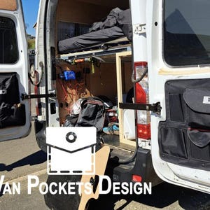 May include: A white van with black fabric storage pockets on the inside of the doors. The pockets are organised with multiple compartments and have red stitching. The van is open and the interior is visible, showing a bed and other items.