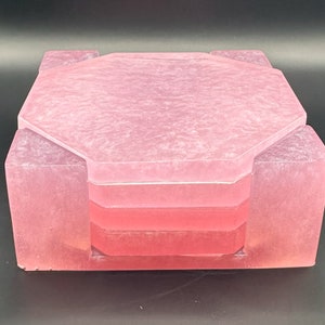 Puede incluir: Un juego de cuatro posavasos octogonales de color rosa con efecto marmoleado. Los posavasos están apilados uno encima del otro.