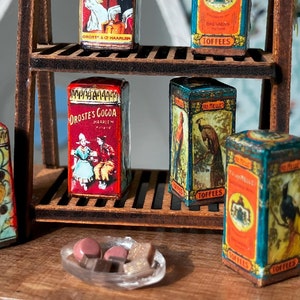 Puede incluir: Una estantería de madera con cinco cajas de hojalata vintage. Las cajas están decoradas con imágenes y texto coloridos. Las cajas están etiquetadas con "Droste's Cocoa" y "Toffels".